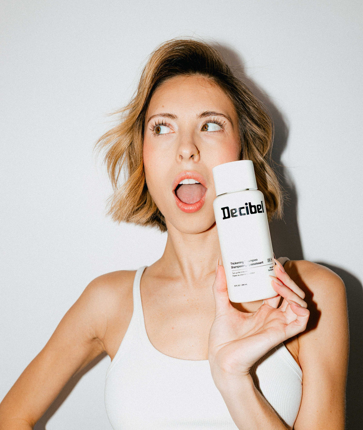 Vrouw met een blonde bob houdt verrast een flacon Thickening Shampoo naast haar gezicht